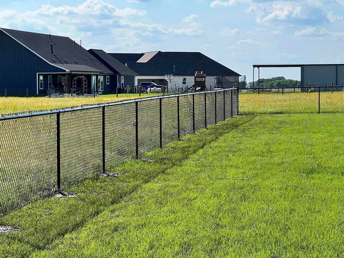 Black metal fence bordering a green field with modern farm buildings in Brookshire TX